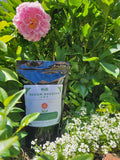 KiS Bloom Booster 2-8-6 pouch standing in a garden between a pink peony bloom and white alyssum.