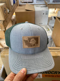 Hand holding a heather gray and green trucker hat with a leather KiS Organics patch