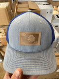 Hand holding a heather gray and royal blue trucker hat with a leather KiS Organics patch